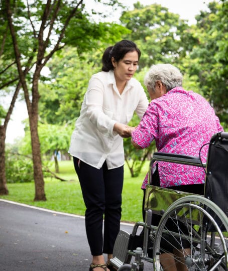 a middle aged woman assisting an elderly woman stand up from a wheel chair in a park surrounded by trees