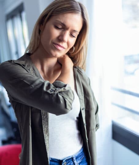 a tired woman standing by the window massaging her shoulder