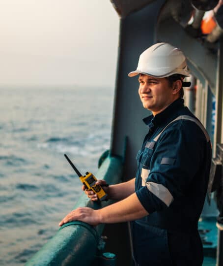 Marine deck worker or officer is shown on deck wearing personal protective equipment holding a walkie-talkie radio with a smile on his face