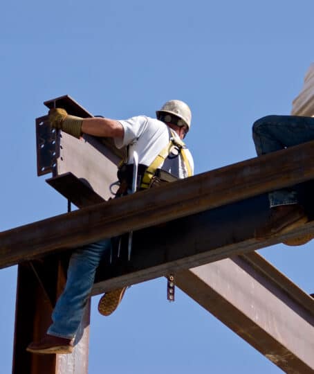 Atop the skeleton of a building, a man is positioning a large beam while the other one observes