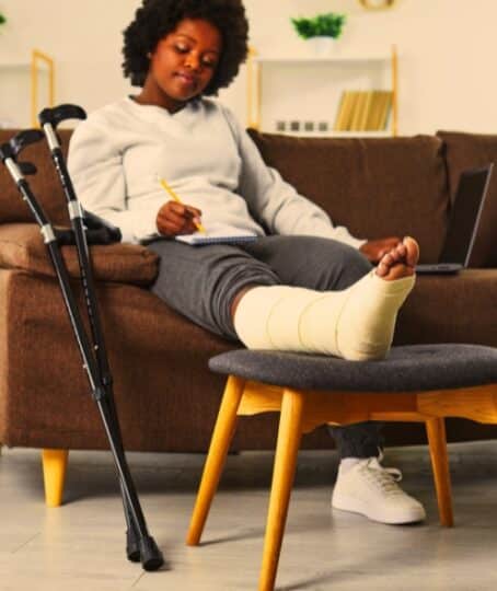 Attractive smiling african american woman works at home in the living room with laptop and makes notes in notebook, sitting on the sofa with broken bandaged leg lying on stool. Crutches are nearby.