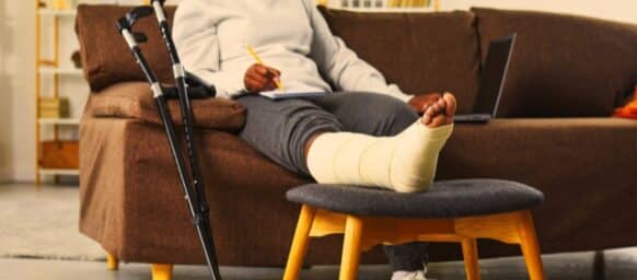 Attractive smiling african american woman works at home in the living room with laptop and makes notes in notebook, sitting on the sofa with broken bandaged leg lying on stool. Crutches are nearby.