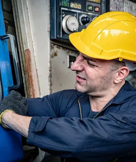 sweaty man with a scrunched up face wearing a hard hat and working gloves tinkering on a machine