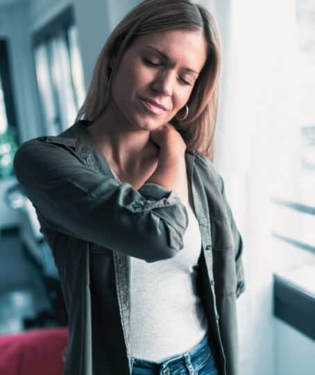 Shot of tired young woman with neck and back pain standing in the living room at home