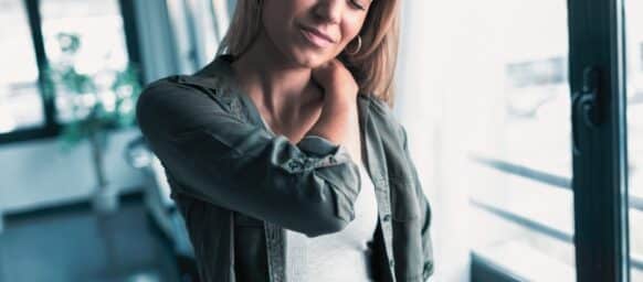 Shot of tired young woman with neck and back pain standing in the living room at home