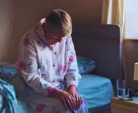 Senior woman sitting on the side of her bed with her head down looking sad.
