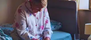 Senior woman sitting on the side of her bed with her head down looking sad.