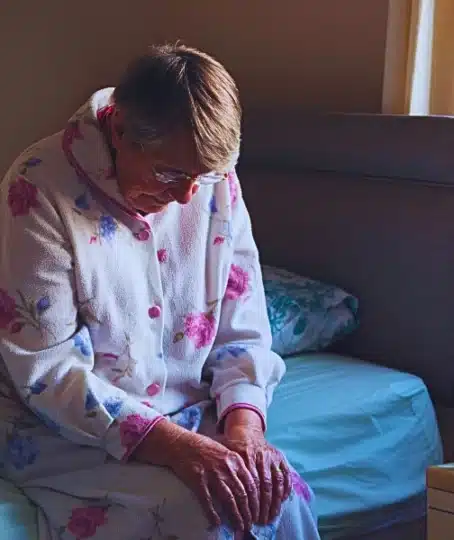Senior woman sitting on the side of her bed with her head down looking sad.
