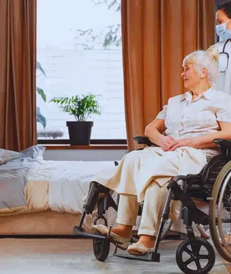 Nurse and an elder woman in a wheelchair inside a room in nursing home. Why Nursing Home Falls Are Still So Common