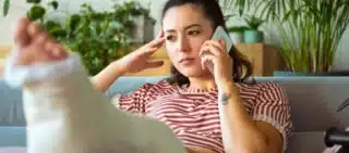 Woman with a leg cast sits on a couch, talking on the phone with a concerned expression.