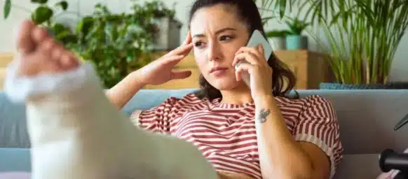 Woman with a leg cast sits on a couch, talking on the phone with a concerned expression.
