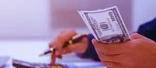 A person holding several US $100 bills in one hand while using a calculator with the other hand. How Average Weekly Wage Is Calculated