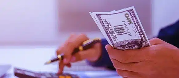 A person holding several US $100 bills in one hand while using a calculator with the other hand. How Average Weekly Wage Is Calculated