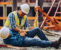 A construction worker sitting on the ground at a construction site, holding their wrist as if injured.