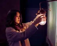 A young woman wearing a light-colored coat, reaching out to hold or adjust a small wall lamp with a warm glowing bulb. Poor Lighting Accidents