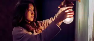 A young woman wearing a light-colored coat, reaching out to hold or adjust a small wall lamp with a warm glowing bulb. Poor Lighting Accidents