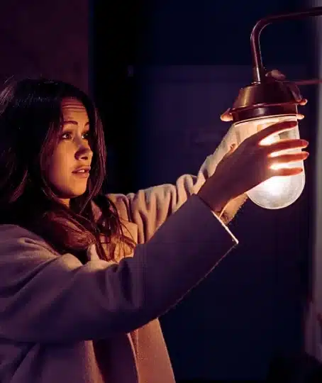 A young woman wearing a light-colored coat, reaching out to hold or adjust a small wall lamp with a warm glowing bulb. Poor Lighting Accidents
