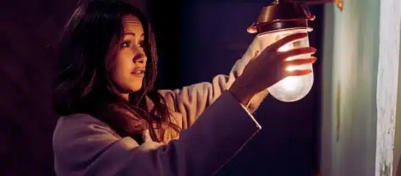 A young woman wearing a light-colored coat, reaching out to hold or adjust a small wall lamp with a warm glowing bulb. Poor Lighting Accidents