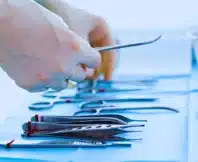 Gloved hands selecting stainless steel surgical instruments from tray.