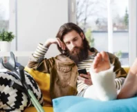 A man with long hair and a beard is lying on a sofa with his leg immobilized and in a cast; he is looking at his cell phone with disappointment, waiting for a response. personal injury claim is delayed