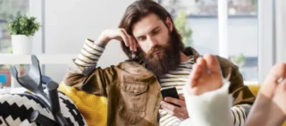 A man with long hair and a beard is lying on a sofa with his leg immobilized and in a cast; he is looking at his cell phone with disappointment, waiting for a response. personal injury claim is delayed