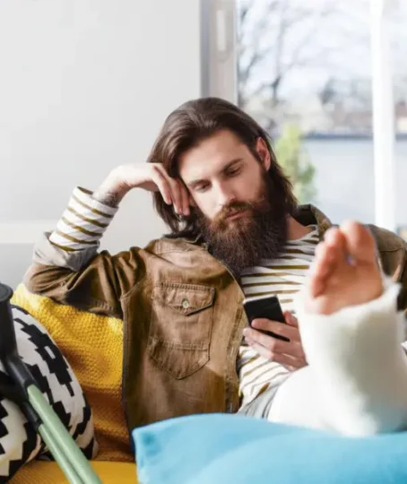 A man with long hair and a beard is lying on a sofa with his leg immobilized and in a cast; he is looking at his cell phone with disappointment, waiting for a response. personal injury claim is delayed