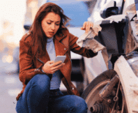 A worried woman looking at her smartphone while crouching next to her damaged car after a traffic accident compensation after a hit-and-run accident, claim for a hit-and-run accident