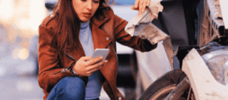 A worried woman looking at her smartphone while crouching next to her damaged car after a traffic accident compensation after a hit-and-run accident, claim for a hit-and-run accident