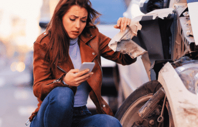 A worried woman looking at her smartphone while crouching next to her damaged car after a traffic accident compensation after a hit-and-run accident, claim for a hit-and-run accident