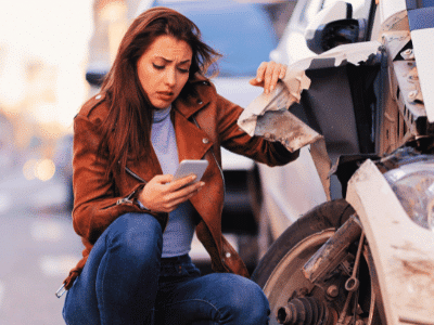 A worried woman looking at her smartphone while crouching next to her damaged car after a traffic accident compensation after a hit-and-run accident, claim for a hit-and-run accident